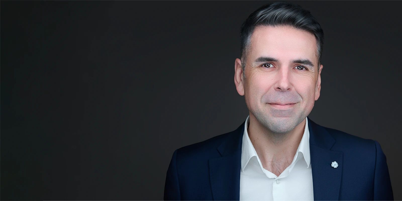 Professional headshot of a man in a navy suit and white shirt on a dark background, with space for headline text.