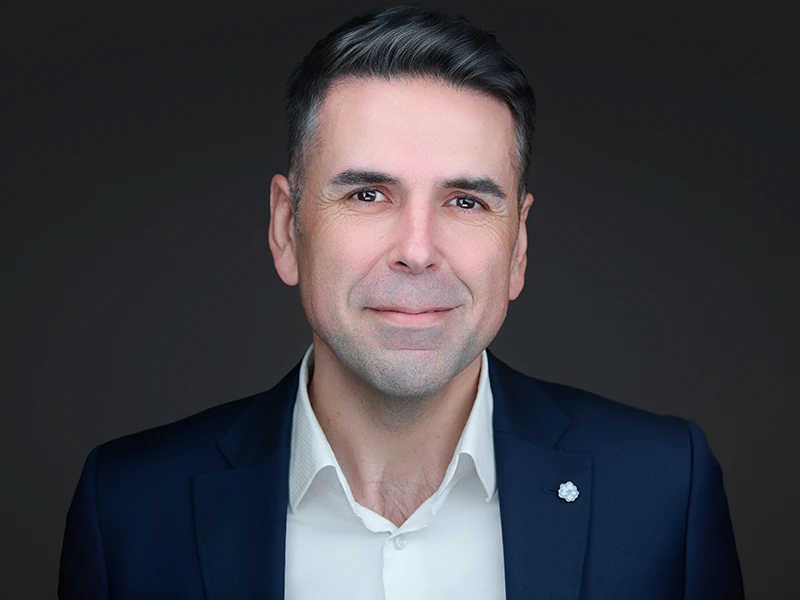 Smiling man with a beard in a light blue shirt, photographed on a dark grey background
