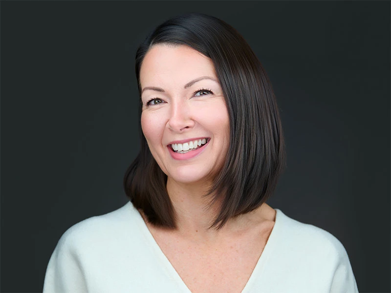 Smiling woman with a dark bob haircut in a white top, photographed on a dark grey background