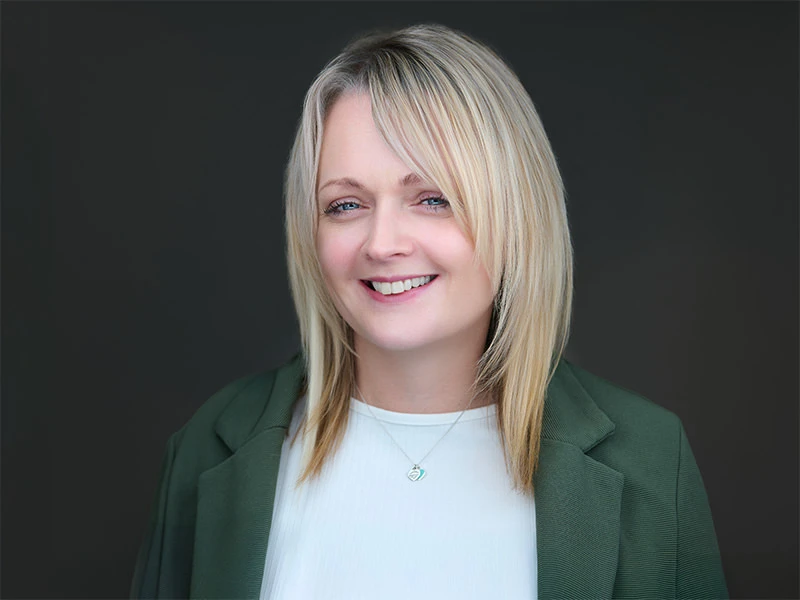 Smiling blonde woman in a green blazer and white top, photographed on a dark grey background