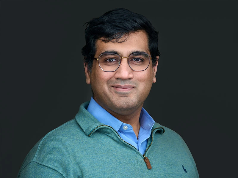 Man wearing glasses in a green quarter-zip jumper and blue shirt, photographed on a dark grey background