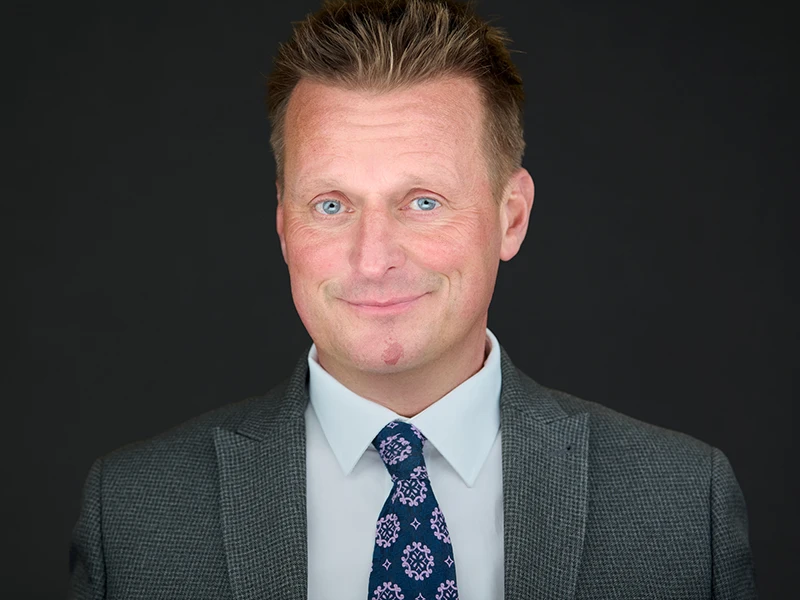 Man in a grey suit with a patterned tie, photographed on a dark grey background