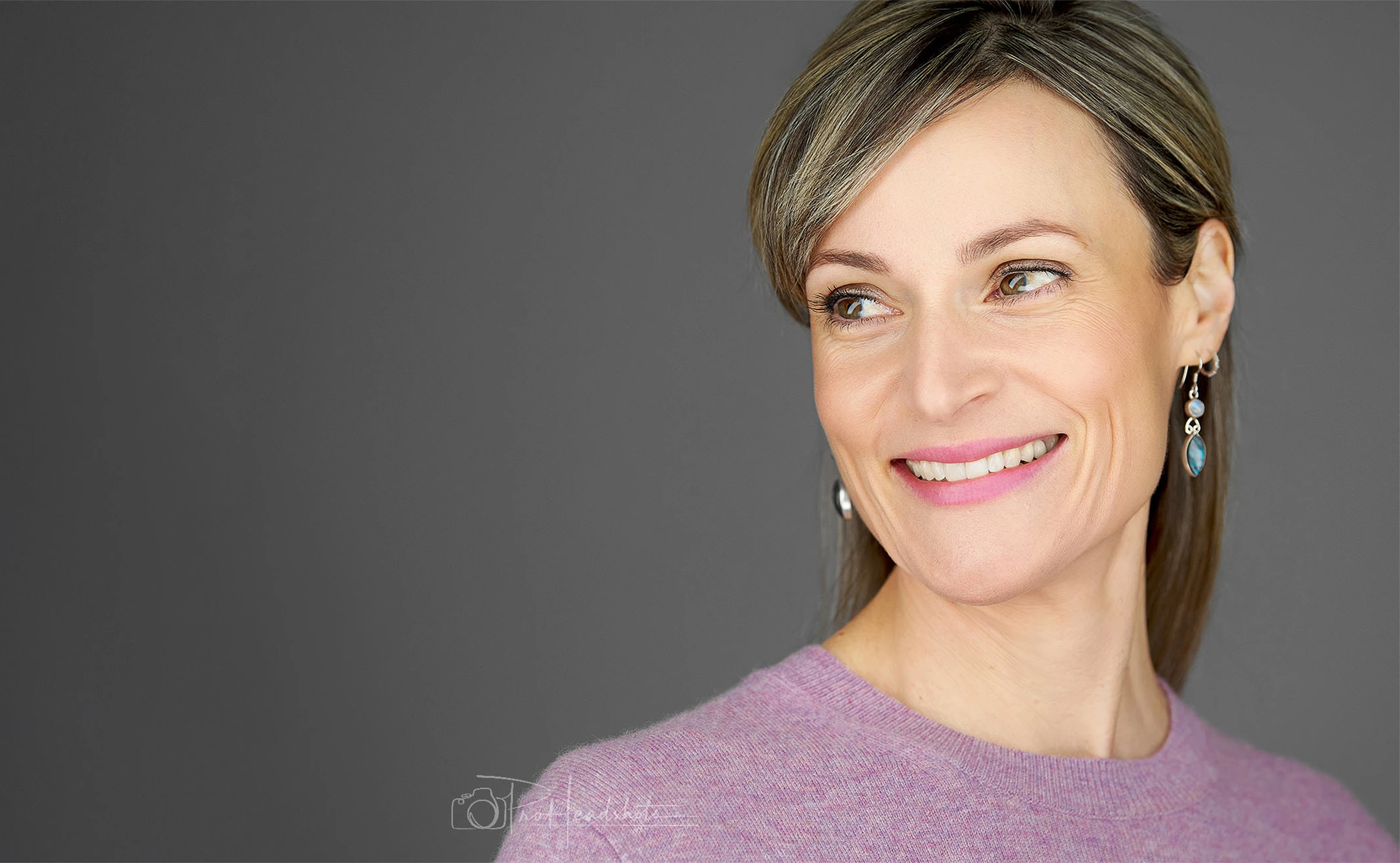 Professional headshot of a woman wearing a mauve sweater and blue drop earrings, showing a natural, engaging smile against a dark background.