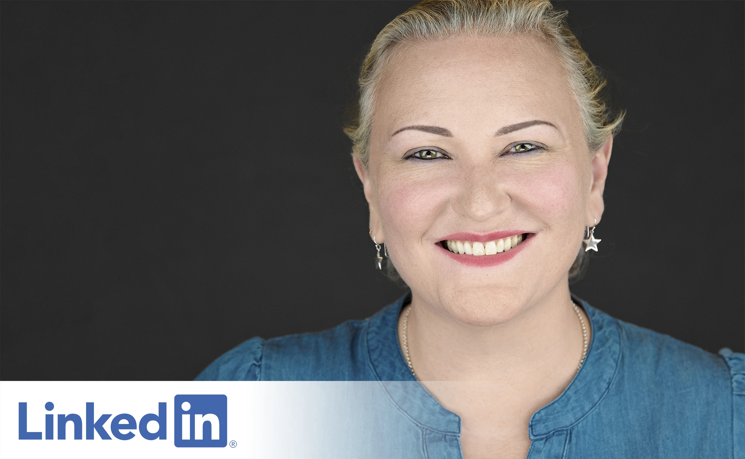 A person with blonde hair wearing a blue top smiles warmly at the camera against a dark background, demonstrating a natural, relaxed expression. Their genuine smile and confident pose show how comfortable they feel being photographed.