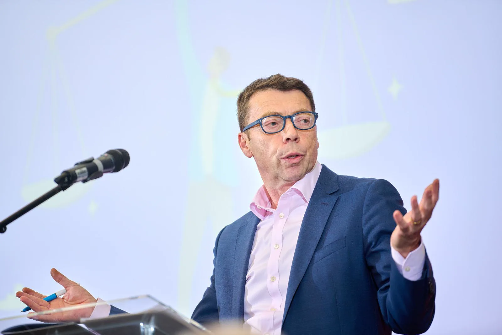 Speaker in blue suit at a clear acrylic lectern, holding a clicker and gesturing naturally with audience visible in the foreground