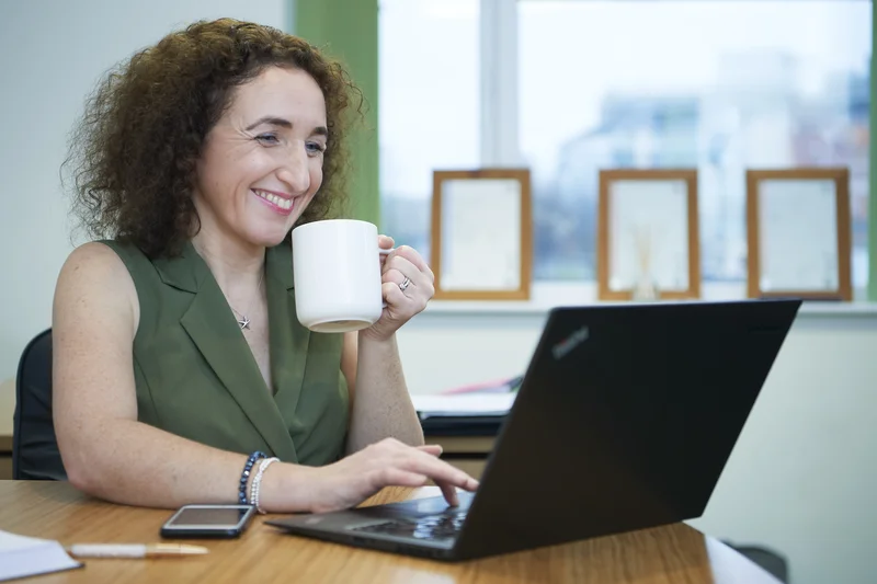 Lizzie Butler smiling at a laptop with a coffee mug, relaxed working mode in a bright office