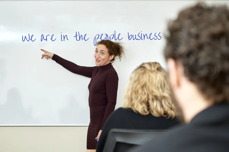 Lizzie Butler facilitating a workshop at a whiteboard reading 'we are in the people business', pointing at the text with energy and confidence