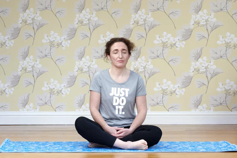 Lizzie Butler seated cross-legged in a yoga meditation pose on a blue mat, eyes closed, against floral wallpaper