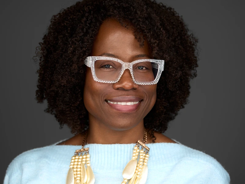 Character-led headshot of a woman with curly hair and white glasses on a dark background