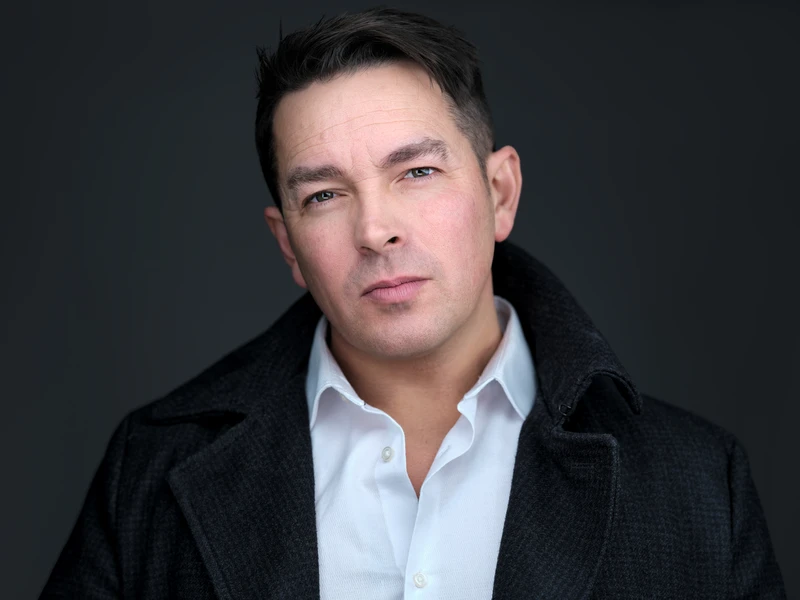 Actor-style headshot of a man in a dark coat on a light background