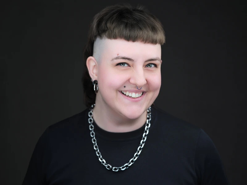 Character headshot of a person with short hair and a chain necklace on a dark background