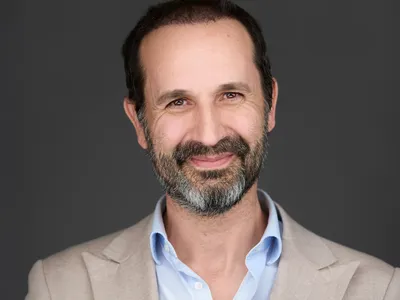 Mature professional with full beard in light grey blazer and blue shirt, confident smile