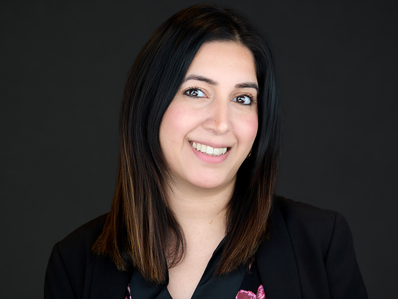Professional corporate headshot of a businesswoman with long dark hair wearing a black blazer against a dark background