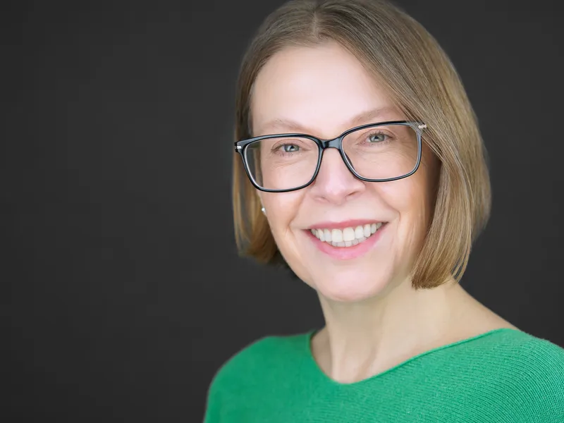Professional woman with bob haircut and glasses in green sweater, smiling confidently against dark background