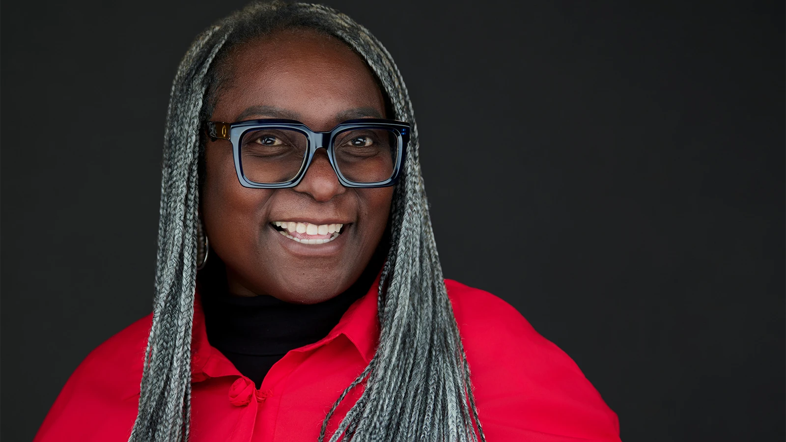 Professional headshot of a woman wearing a mauve sweater and blue drop earrings, showing a natural, engaging smile against a dark background.