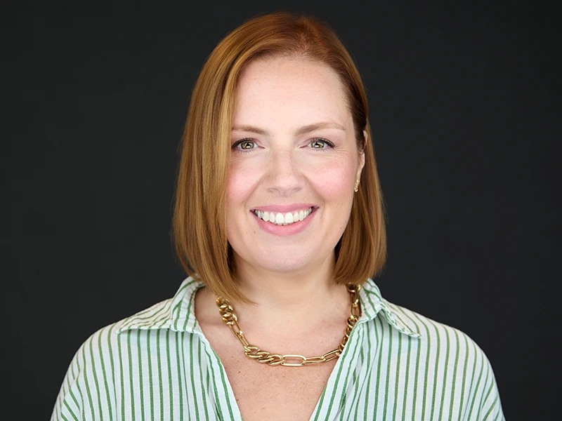 Smiling female professional with red hair, green striped shirt and gold chain on a dark background, London headshot
