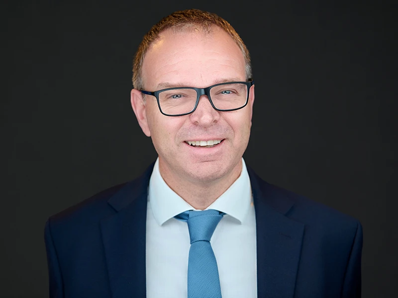 Smiling male professional wearing glasses, navy suit and light blue tie on a dark background, London headshot