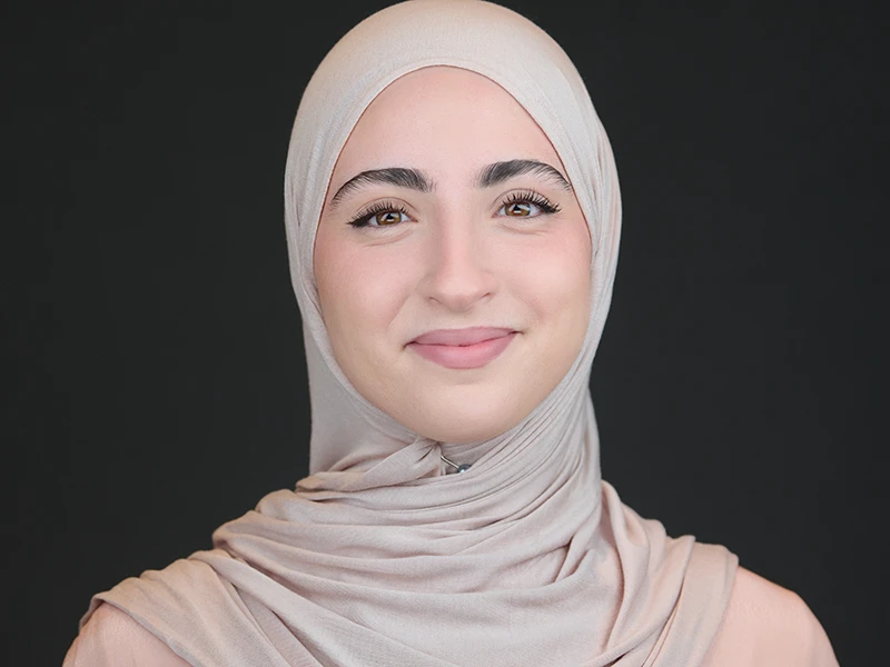 Professional portrait of a woman wearing a light beige hijab, natural smile, dark studio background