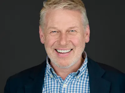 Professional headshot of businessman in navy blazer and blue gingham shirt, smiling warmly against dark background