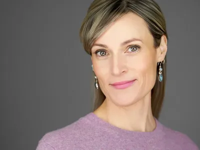 Confident professional woman in lilac jumper with drop earrings, smiling subtly against a neutral background, business headshot.