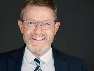 Professional headshot of a business executive in navy suit and striped tie wearing glasses, smiling against a dark background.