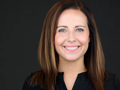 Smiling woman with straight brown hair in black top, professional headshot on dark background.