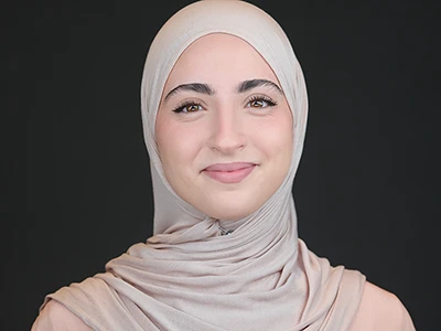 Smiling young woman wearing a light beige hijab, professional headshot on dark background.