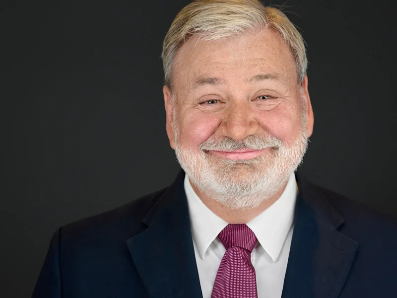 Company chairman with distinguished white beard in classic navy suit and burgundy tie.