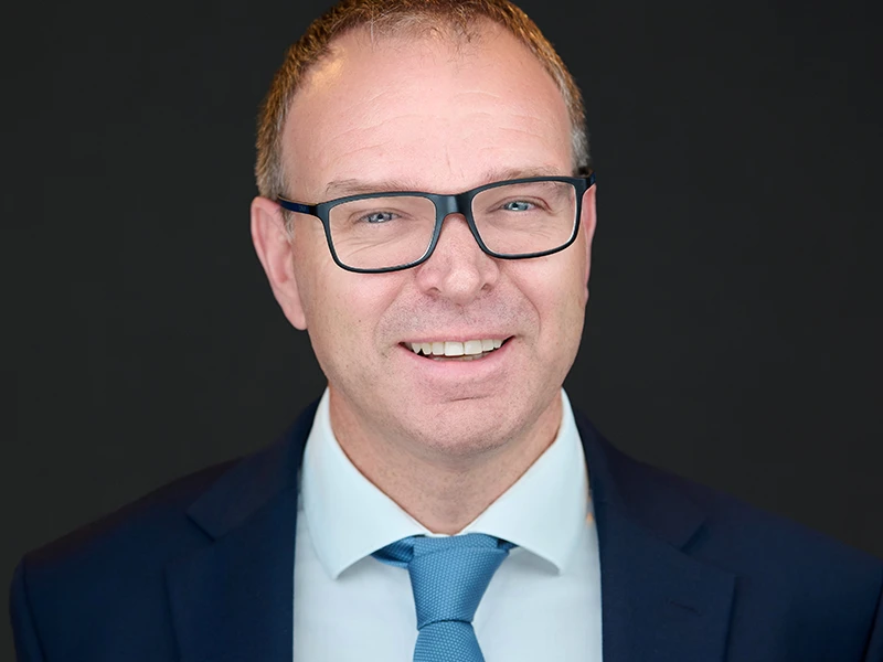 Smiling professional man in glasses wearing a navy suit and light blue tie, photographed on a dark background.
