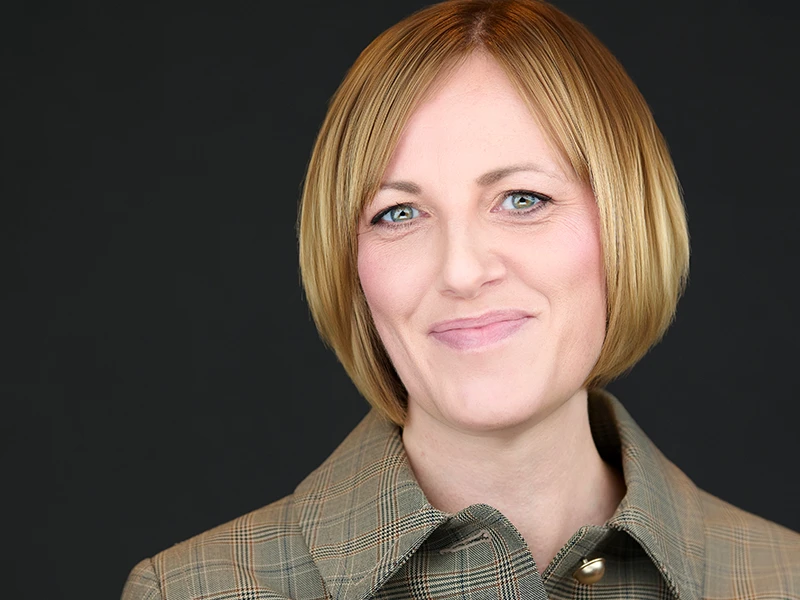 Smiling professional woman with short blonde bob wearing a green checked jacket, photographed on a dark background.
