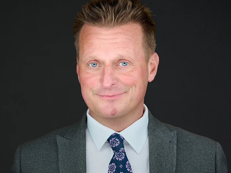 Professional headshot of a man in a grey suit and patterned tie, smiling confidently against a dark background.