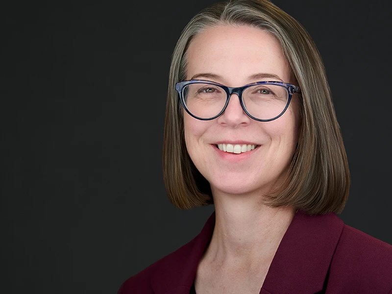 Professional headshot of a woman wearing glasses and a burgundy jacket, smiling warmly against a dark background.