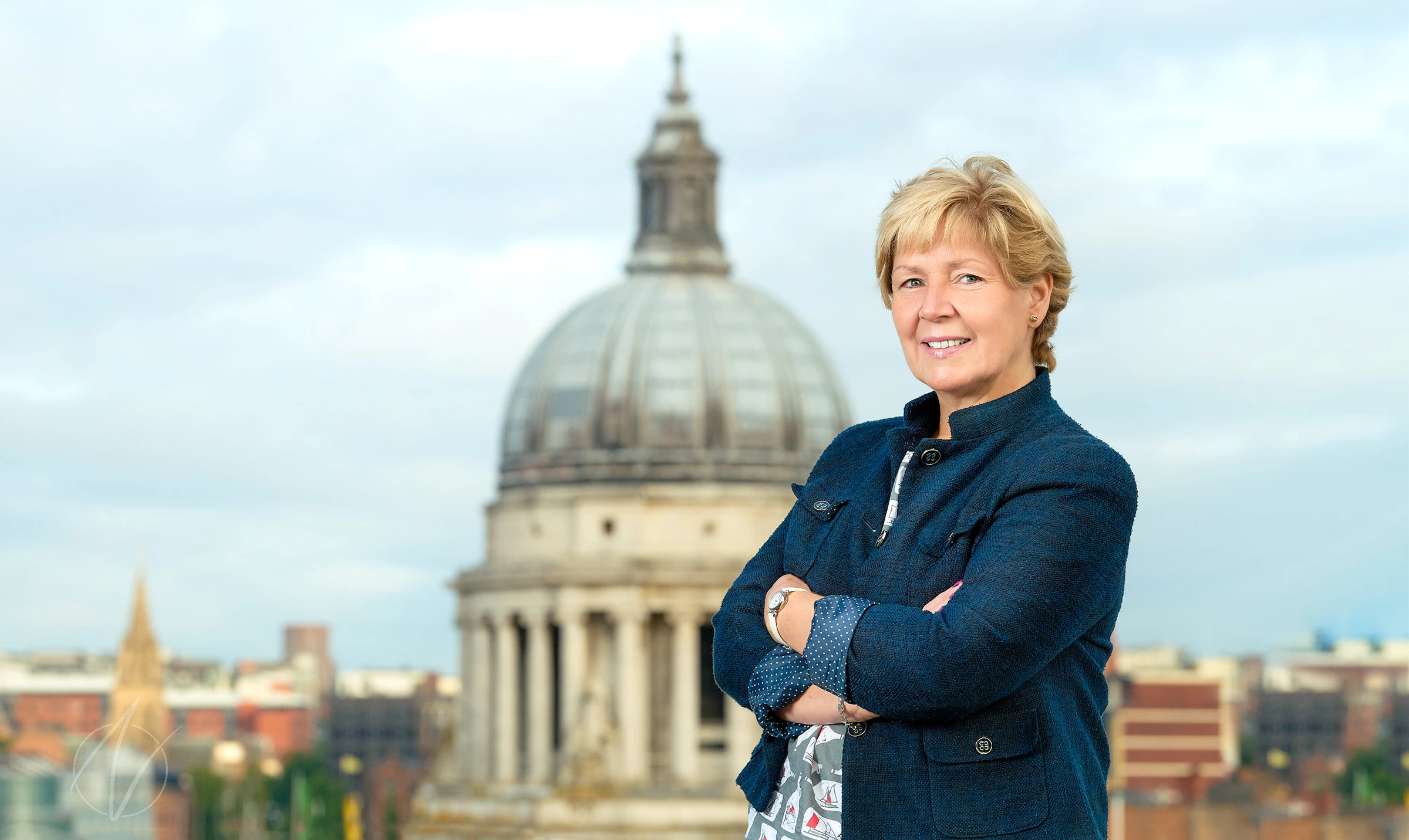 A confident business leader stands with arms crossed in a navy jacket, photographed against an impressive cathedral and city skyline backdrop, adding gravitas to this executive portrait.