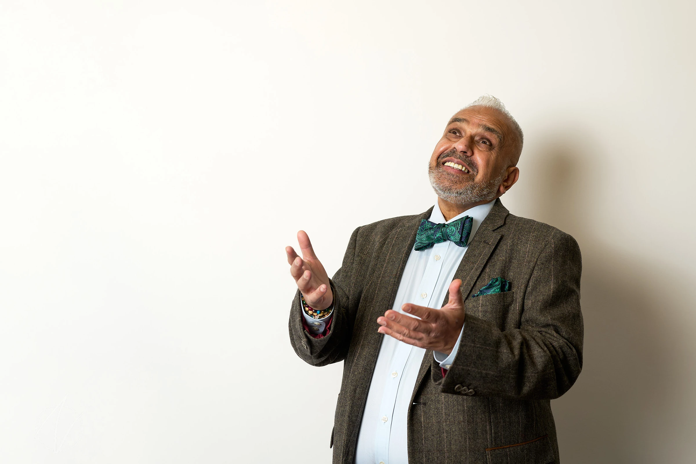 A distinguished man in a grey pinstriped suit with teal bow tie gestures animatedly with raised hands, his genuine smile and dynamic pose conveying enthusiasm and expertise against a clean white background.