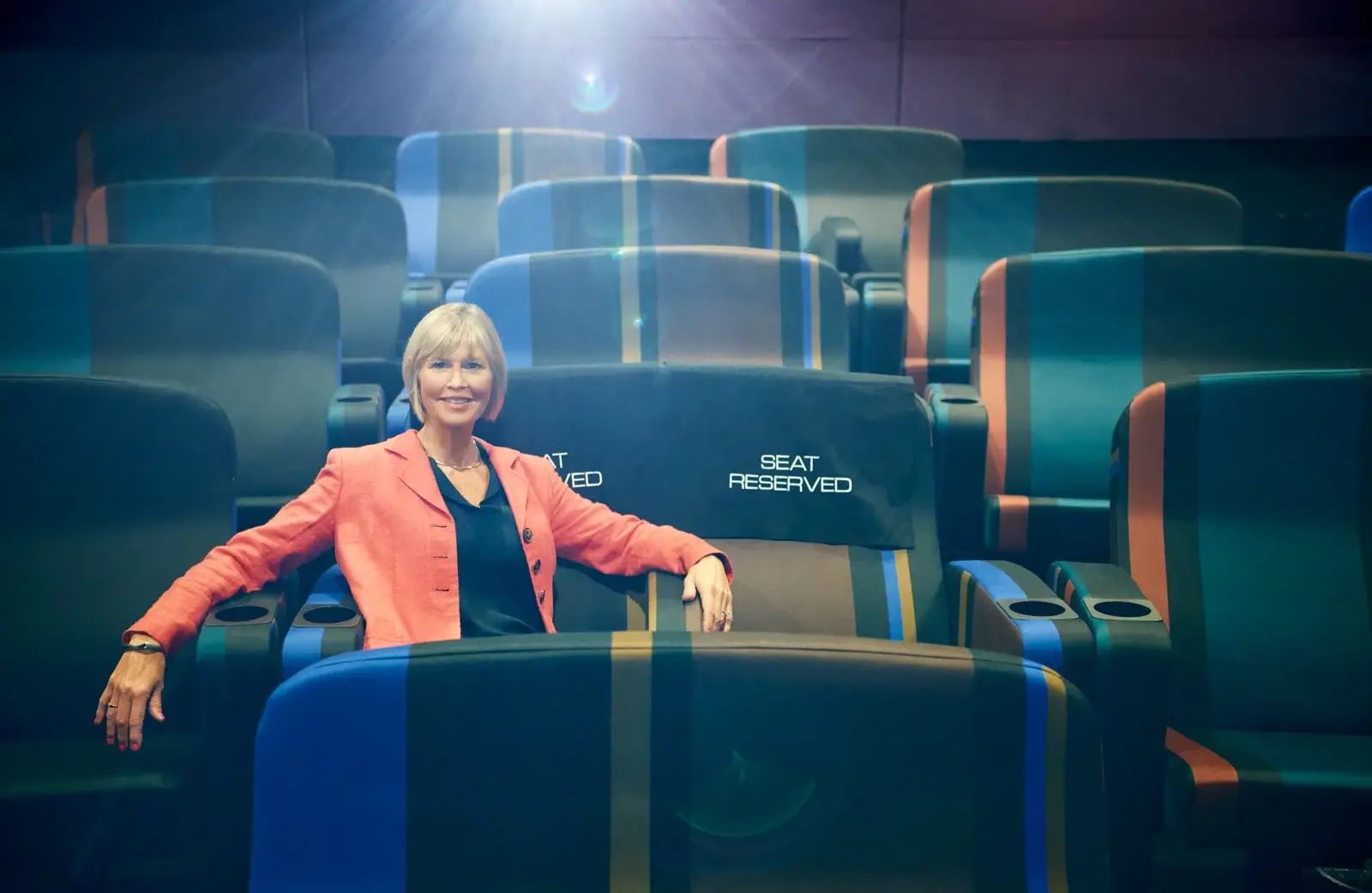 A confident female speaker in a coral blazer sits among modern theatre seating, showcasing executive presence and thought leadership in a professional setting.