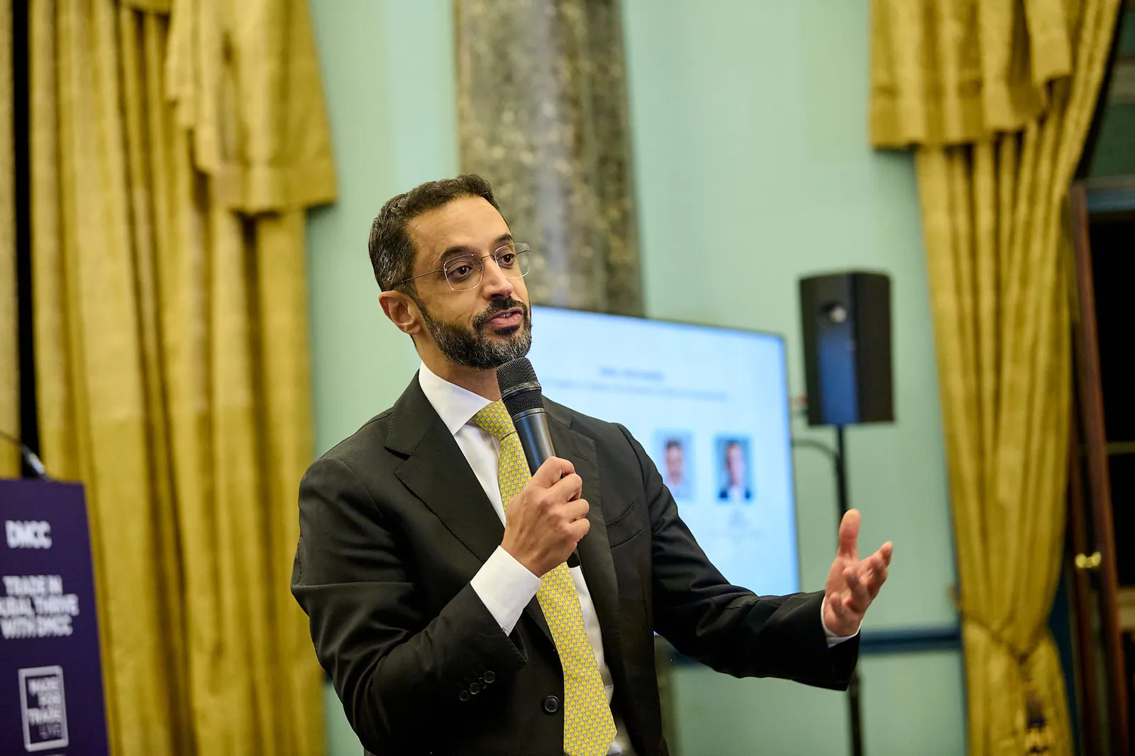 A speaker with a handheld microphone during the panel at Made for Trade Live London, gold curtains and a screen behind him