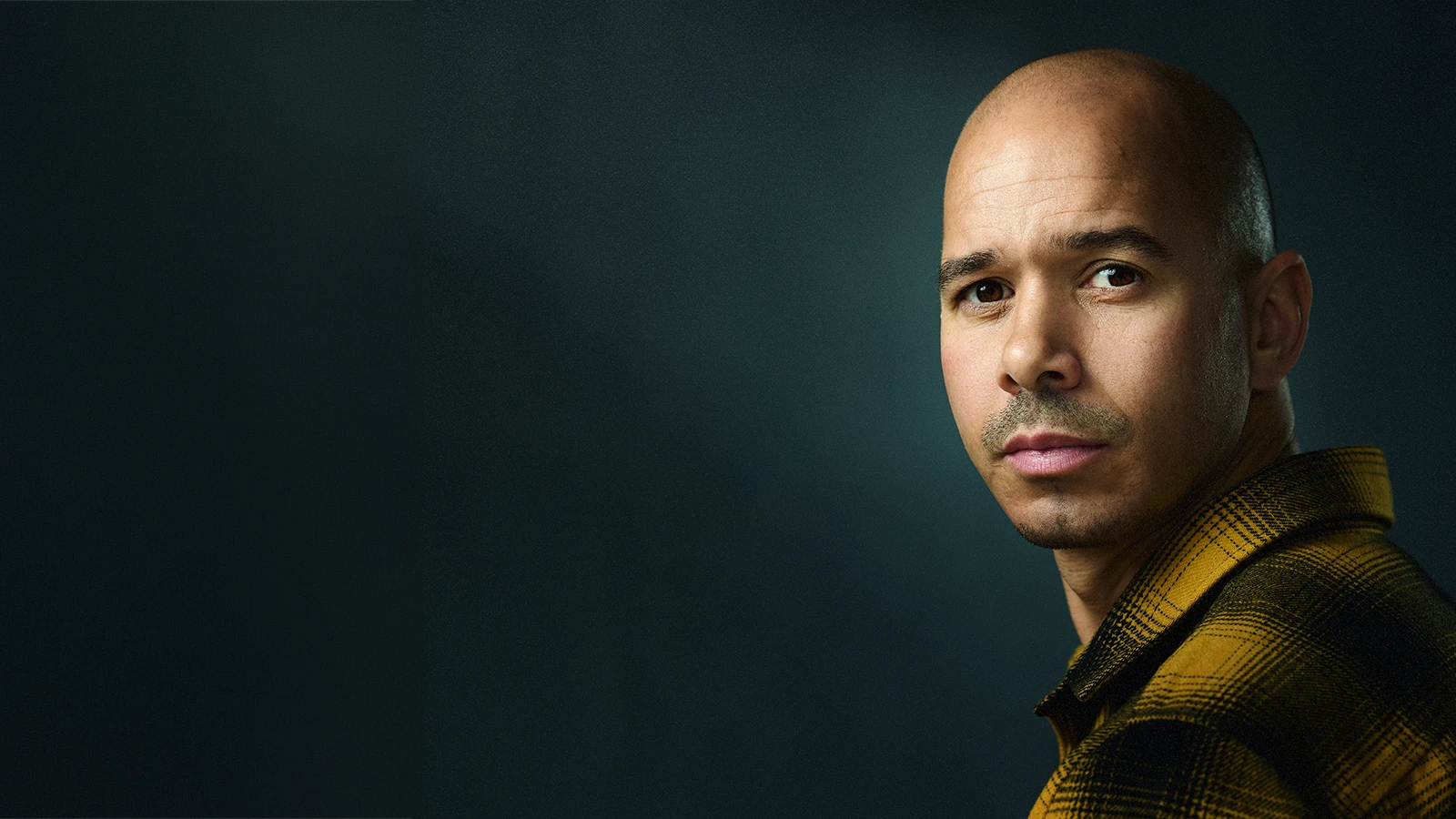 A statement headshot with a dark background and dramatic light, the subject looking over his shoulder straight down the lens