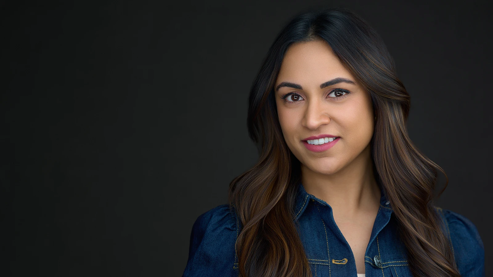 Woman with long dark wavy hair and warm smile in a navy denim jacket, professional headshot by a Cheltenham and Gloucester headshot photographer