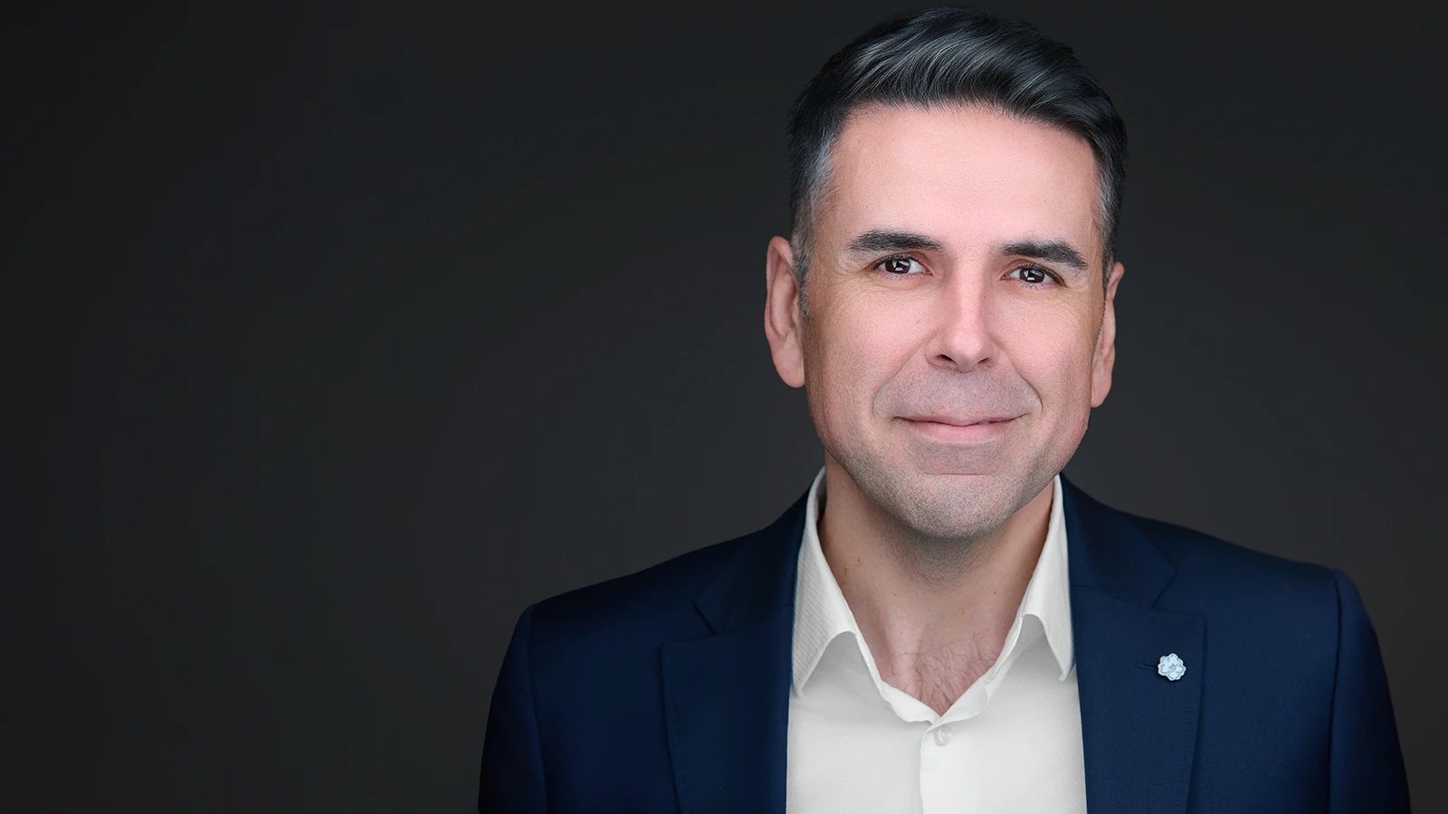 Professional headshot of a man in a navy blazer and open-collar cream shirt, smiling against a dark studio background — taken on location in Cornwall