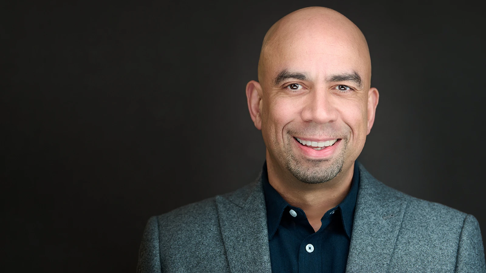 Professional man with shaved head wearing a grey blazer and navy shirt, smiling confidently against a dark background — Plymouth headshot photography by ProHeadshots.