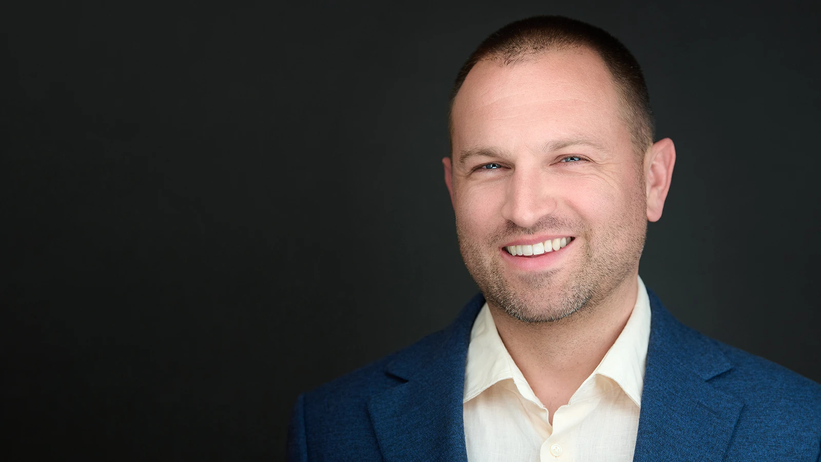 Professional headshot of a man in a blue blazer and cream shirt, smiling against a dark studio background — taken on location in Somerset