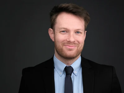 Bearded man in a dark suit looking composed and polished in a studio headshot