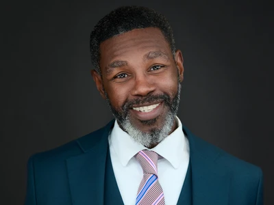 A bearded man in a dark suit, strong and composed expression against a studio background