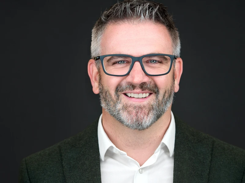 Headshot of a bearded man in glasses and a blazer on a dark studio background