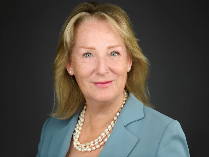 Executive corporate headshot of a woman in a pale blue blazer and pearl necklace, warm approachable expression, dark studio background