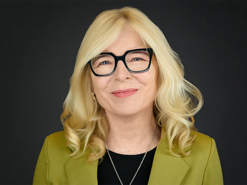 Headshot of a woman in glasses, wearing a blazer on a dark studio background