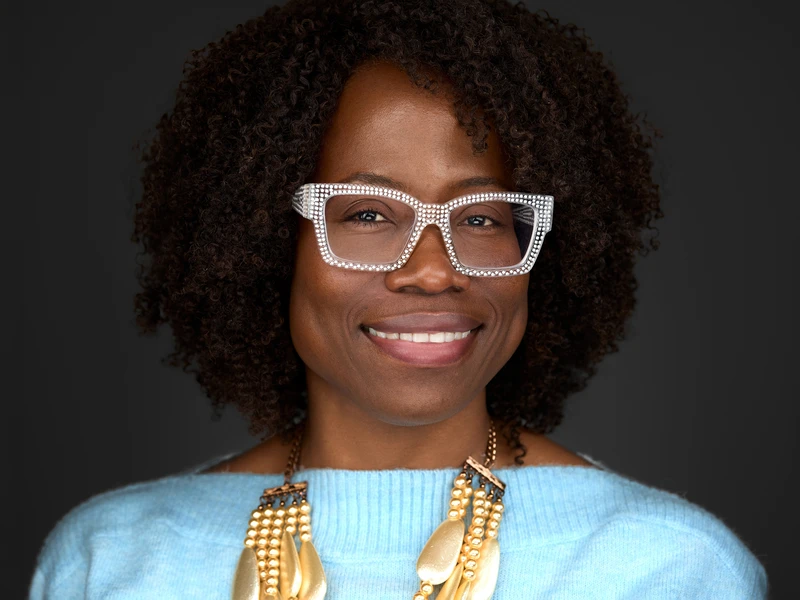 Corporate headshot of a woman in statement glasses and a pale blue jumper, warm confident smile, dark studio background