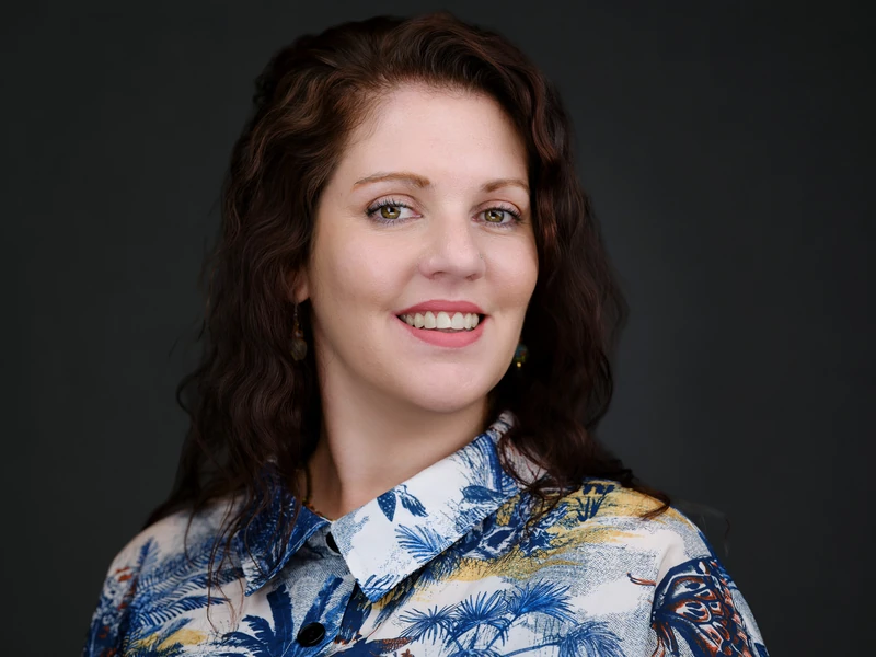Professional studio portrait of a woman with wavy brown hair in a patterned blouse on a dark grey background